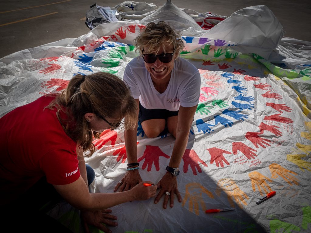 Wendy Tuck is centre frame, with Erica Lush drawing around her hands in an orange pen. Wendy is sitting in the middle of the handprints and smiling, wearing sunglasses.