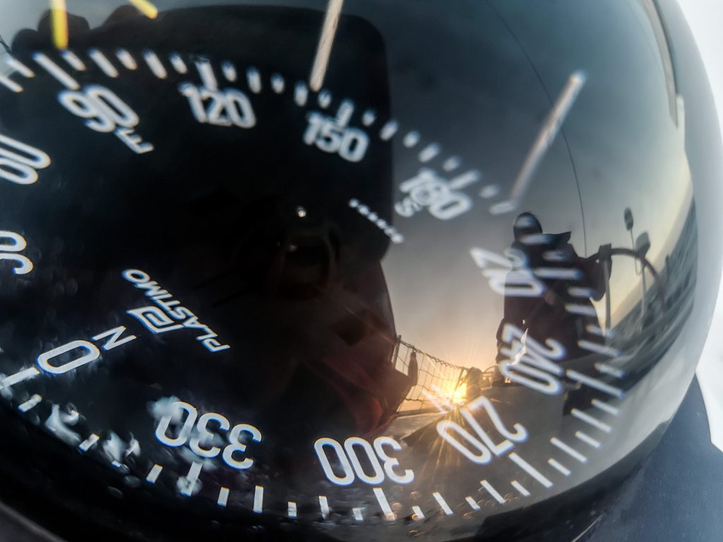 A compass, reflecting Maiden's skipper Wendy Tuck, who is at Maiden's helm. There is a sunset in the background