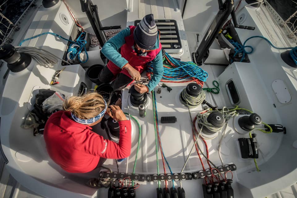 Two sailors sit on the deck of the yacht Maiden. They are both working on winches. The deck has a lot of ropes everywhere, and it is clear there is a lot of work going on. 
