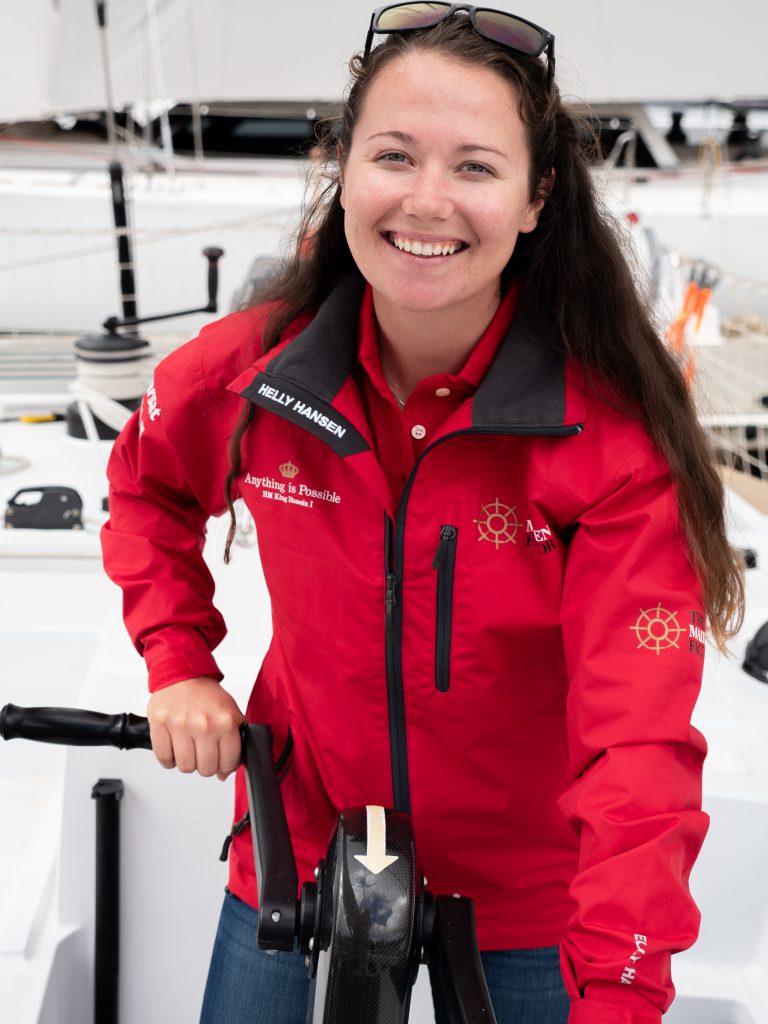 Alison stands at the grinder, with both hands on either side. She had sunglasses on her head and is smiling. She wears a red Maiden jacket with a red polo underneath. Her hair is down.