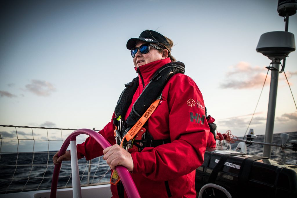 Kate Palfrey stands at the wheel of Maiden, in the centre of the image. She is wearing a red jacket and a black lifejacket. She is wearing sunglasses, reflecting the ocean, and wearing a black cap. In the background, the water is dark, and the sky is bright.

