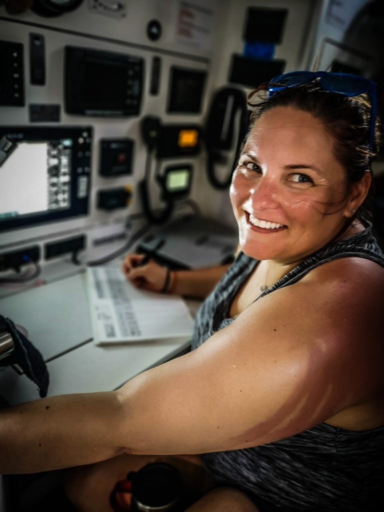 Down below deck, a sailor is writing and smiling at the camera. The nav station is behind, with lots of equipment.