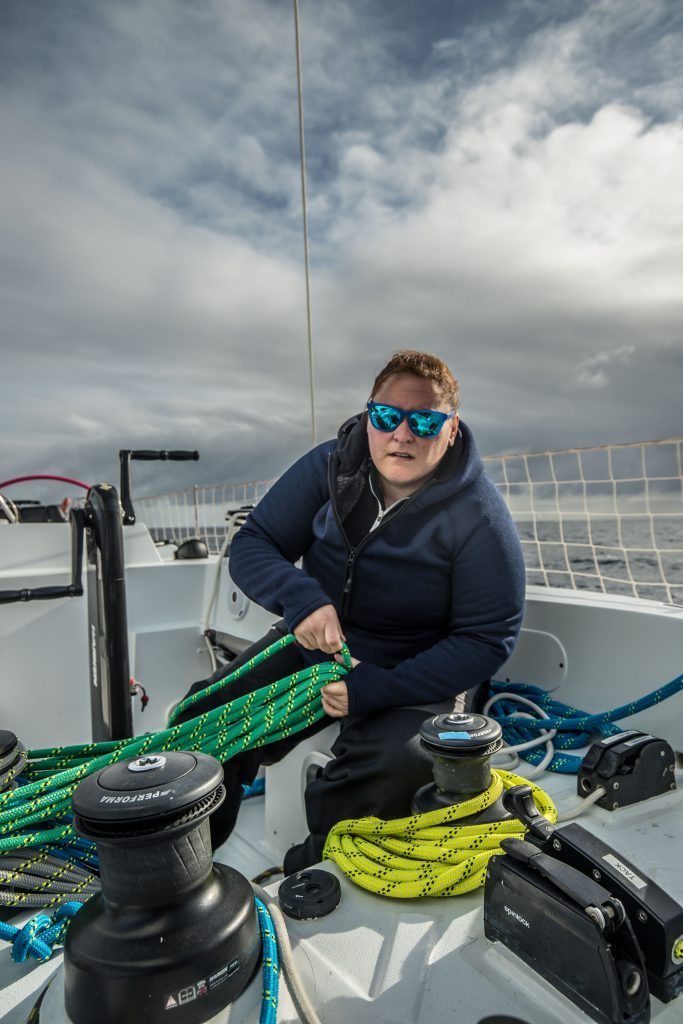 A sailors on Maiden is working with ropes on deck. She wears reflective blue sunglasses and warm clothing.