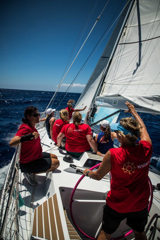 The crew sit on Maiden, photo taken from the back of the boat, the boat at a slight angle. The ocean is deep blue in front and the sky light blue with a few clouds. 