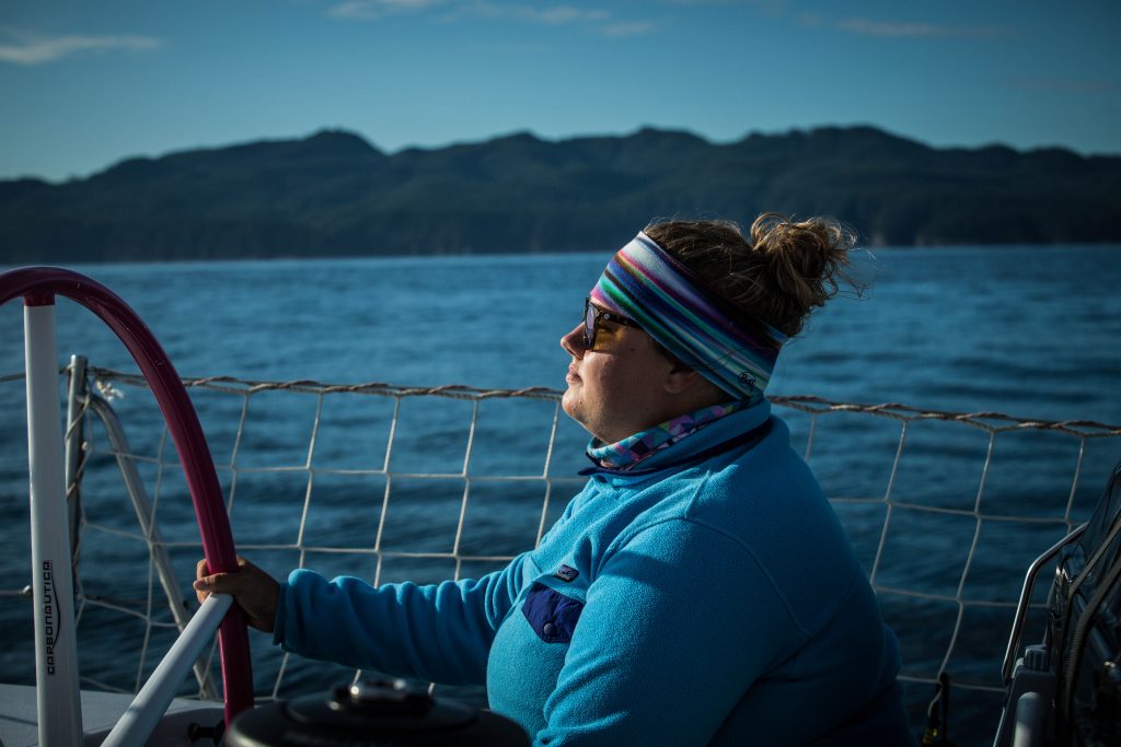 Kirsten at the helm, taken side on. She wears blue, wears glasses and a headband and holds the wheel. Islands are in the background 